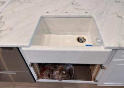 White utility sink on a marble countertop; open cabinet beneath reveals a trash bag and plumbing.
