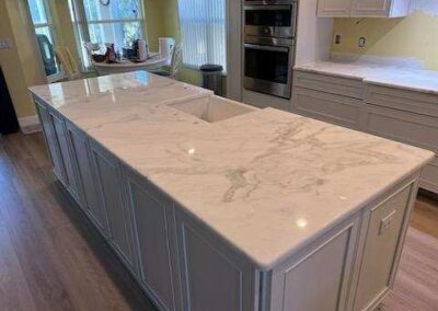 White kitchen island with a marble countertop and sink in a bright, modern kitchen; white cabinets and stainless appliances in the background.