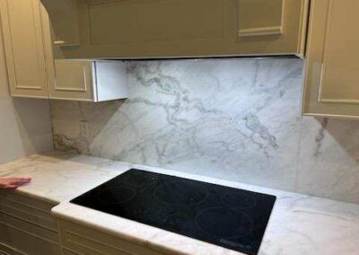 White kitchen with marble backsplash and counters, a black glass‑ceramic electric cooktop, and cream cabinets above and below.