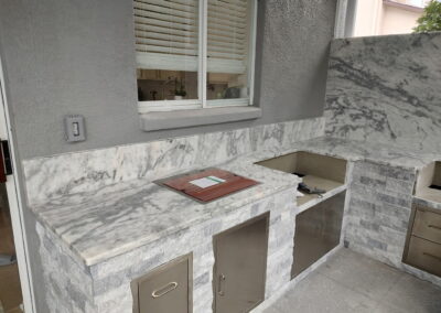 Outdoor kitchen area with marble countertops and built-in stainless steel cabinets along a gray stucco wall, a window with blinds behind.