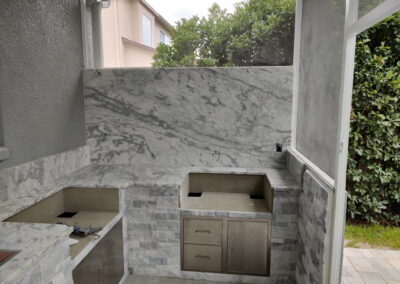 Outdoor marble-covered barbecue area with stainless steel drawers and counters, surrounded by stone walls and green hedges