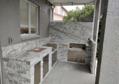 Outdoor kitchen area with marble countertops, gray stone cabinets, built-in grill, and string lights under a covered patio.